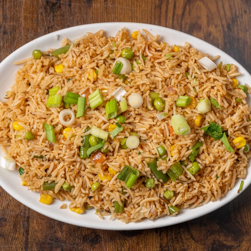 A white oval plate filled with Indian fried rice, garnished with green onions, peas, and corn.