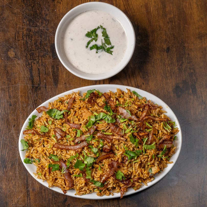 Indian biryani with fried onions and cilantro, served with a side of raita on a wooden table.