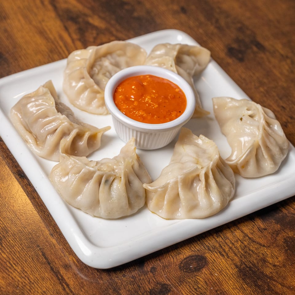 A white rectangular plate of Indian momos, with a small bowl of red dipping sauce in the center.