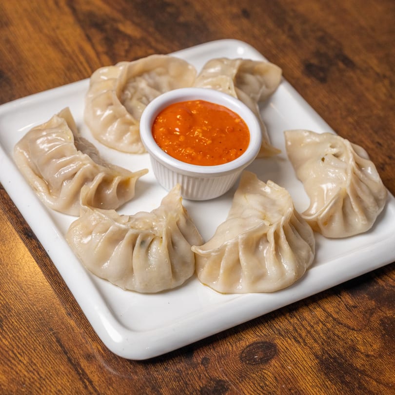 A white rectangular plate of Indian momos, with a small bowl of red dipping sauce in the center.