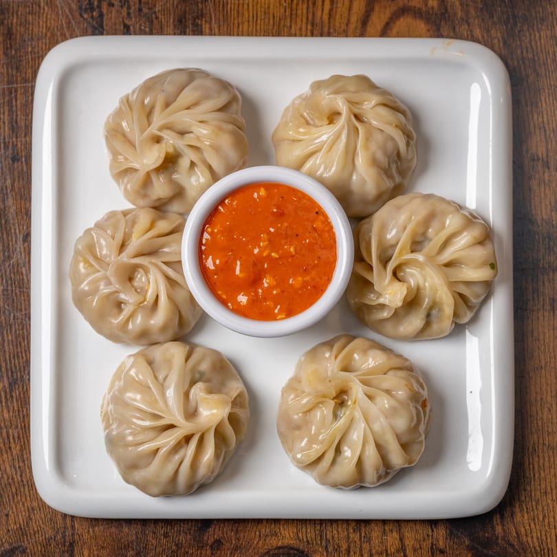 Six Indian momos, steamed dumplings, arranged around a small bowl of red chili sauce on a white plate.