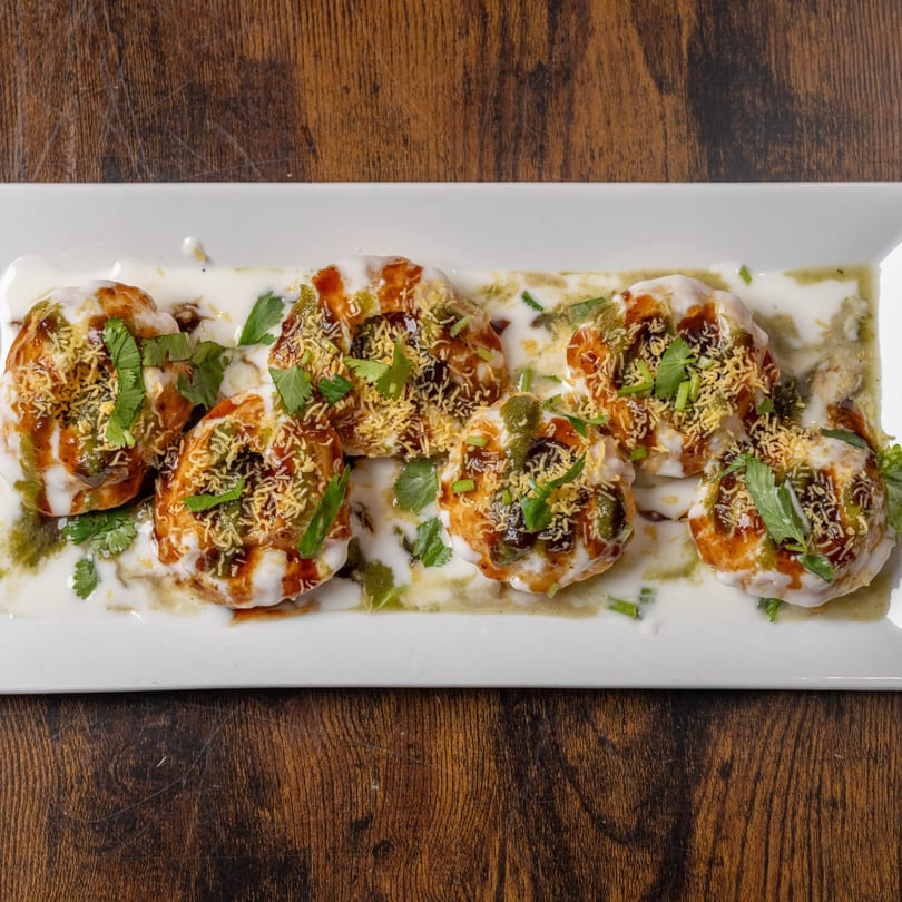 Six Indian Delight Dahi Puri, topped with yogurt, chutneys, cilantro, and sev, on a white plate.